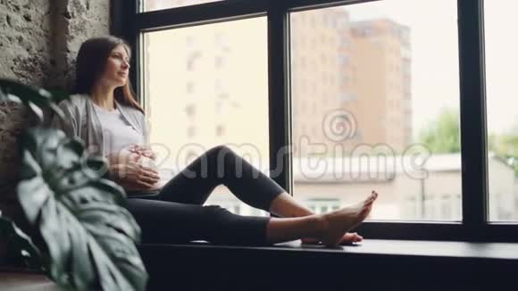 年轻漂亮的准妈妈坐在窗台上抚摸着肚子从窗外欣赏风景视频的预览图