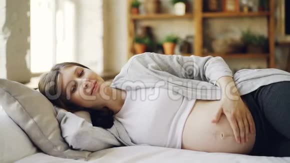 美丽的年轻女子期待着母亲躺在家里的床上抚摸着她的腹部表达对她孩子的爱不错视频的预览图