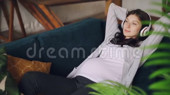 怀孕的年轻女士正在听音乐无线耳机放在家里客厅的沙发上美丽美丽视频的预览图