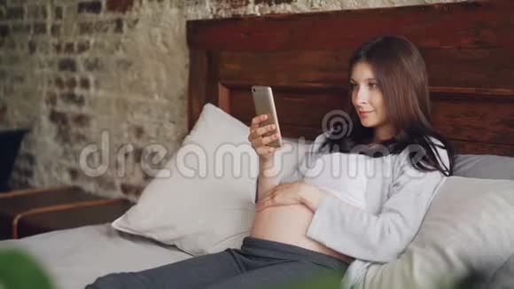 漂亮的年轻女人期待妈妈正在使用智能手机抚摸她的腹部微笑地躺在家里的床上现代视频的预览图