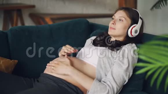 快乐的孕妇躺在沙发上戴着耳机听音乐然后在肚子上戴上耳机视频的预览图