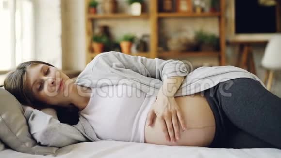 迷人的孕妇躺在床上休息用关心和爱抚她的婴儿腹部可爱的母亲视频的预览图