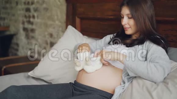 怀孕的年轻女士手里拿着可爱的婴儿鞋在肚子上做小动作与未出生的孩子玩耍视频的预览图