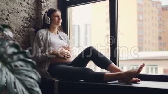 漂亮的孕妇正在抚摸她的腹部在家坐在窗台上听着耳机里的音乐视频的预览图