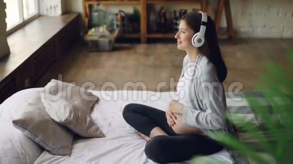 快乐的期待妈妈抚摸她的肚子通过耳机听音乐躺在舒适的双人床上视频的预览图