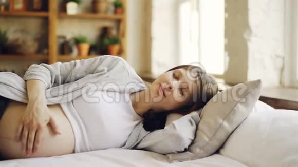 怀孕的女孩躺在床上闭着眼睛用温柔和爱抚摸她的腹部健康怀孕视频的预览图