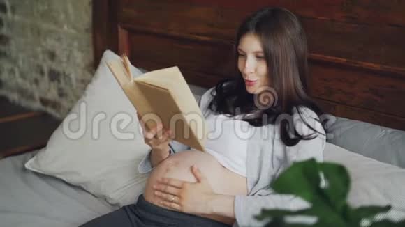 微笑的女孩期待着母亲大声朗读书给她未出生的婴儿微笑着躺在现代公寓的床上视频的预览图