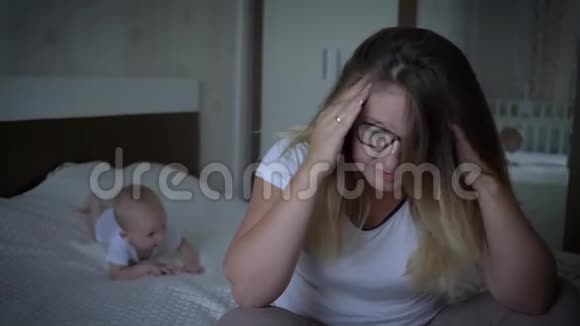 心烦意乱的年轻妈妈坐在床的边缘在家里爬行的婴儿的背景下视频的预览图