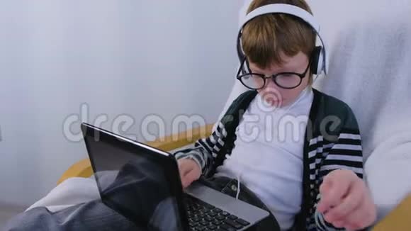 男孩用耳机和坐在扶手椅上的眼镜在笔记本电脑上写和键入信息视频的预览图