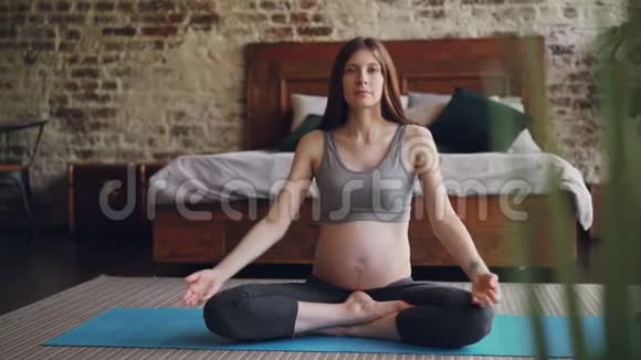 好看的怀孕少女正坐在家里坐着莲花姿势坐在瑜伽垫上举起手臂然后把手掌放在上面视频的预览图