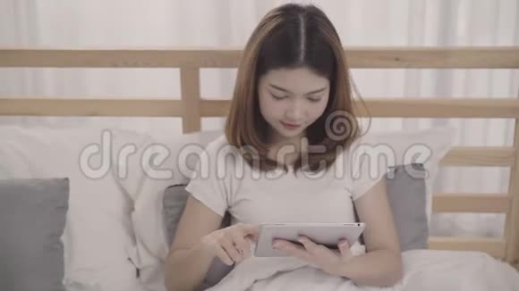 年轻的亚洲女人在早上醒来后躺在床上使用平板电脑视频的预览图