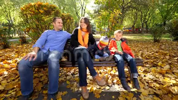 美丽的秋天公园里有两个孩子坐在长凳上的家庭画像视频的预览图