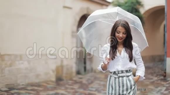 微笑着年轻的黑发女人穿着衣服带着雨伞沿着一个古老的城镇的街道散步在雨下散步视频的预览图