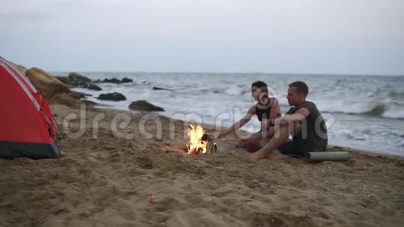 一对年轻夫妇的慢镜头男人和女人坐在空荡荡的海滩上树贩们正在烤香肠视频的预览图