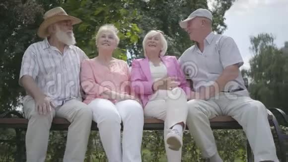 两对可爱的成熟夫妇在夏天公园里坐在长凳上聊天和微笑老年夫妇双人约会视频的预览图