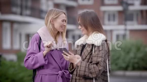 两个女孩带着智能手机在秋天在户外笑女孩穿着时髦的衣服视频的预览图