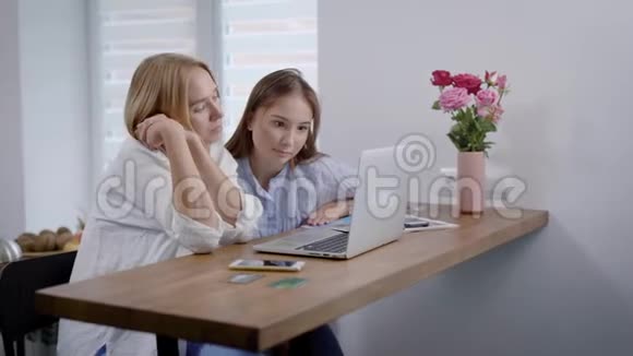 金发和黑发女人坐在厨房的桌子上手提电脑在网上看有趣的视频有趣的室内视频的预览图