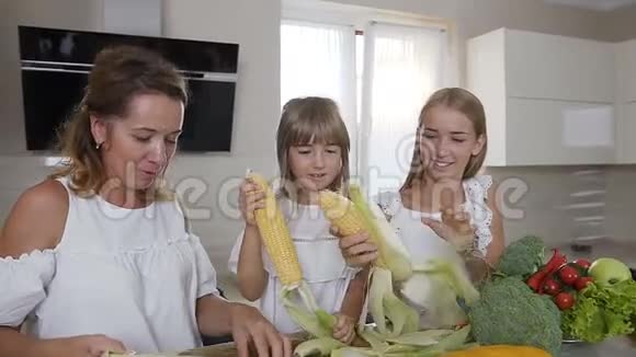 漂亮的妈妈和她的女儿在家里的厨房里从叶子上清洗成熟的玉米耳朵幸福的家庭在打扫卫生视频的预览图