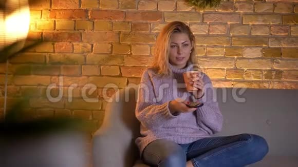 成年白种人金发女性在室内坐在沙发上看电视时捧着一杯热茶的特写镜头视频的预览图