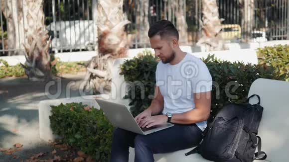 微笑的年轻人在笔记本电脑上工作坐在长凳上拿着旅行箱和包背包游客在度假视频的预览图