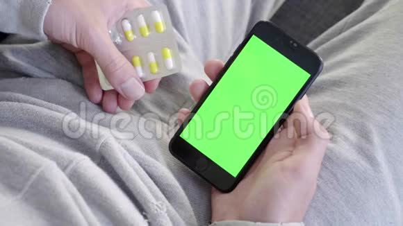 男人在家坐在沙发上拿着绿色的模拟屏幕智能手机吃药视频的预览图