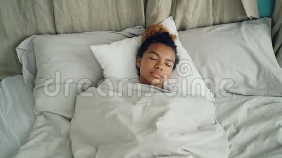 年轻的非洲裔美国妇女睡在舒适的床上在温暖的毯子下休息在美丽的亚麻布上舒适舒适视频的预览图