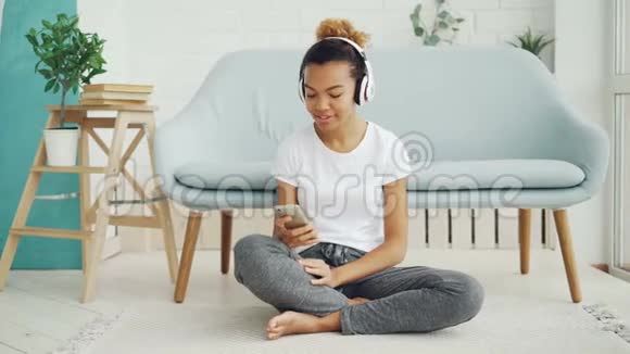 漂亮的女孩正在智能手机上玩游戏一边触摸屏幕一边微笑一边坐着耳机听音乐视频的预览图