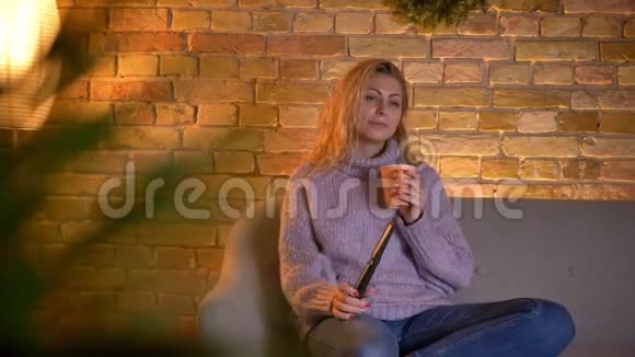 成年白种人金发女性坐在沙发上看电视时端着一杯热茶的特写镜头视频的预览图