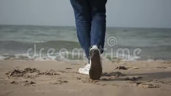 一个穿着米色外套和蓝色牛仔裤的年轻女人在海边跳舞沿着海岸散步视频的预览图