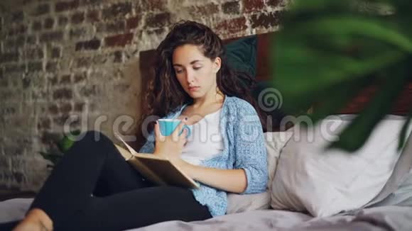 漂亮的年轻女士正在床上看书拿着一杯茶躺在枕头上在家享受一天漂亮的阁楼视频的预览图