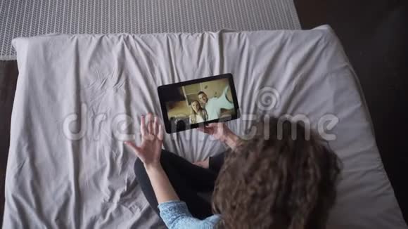 这位年轻的女士在网上和一对漂亮的夫妇聊天平板电脑看着屏幕坐在家里的床上聊天视频的预览图
