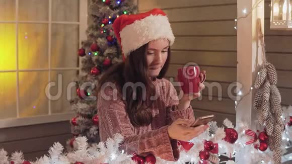 圣诞老人帽子里的布鲁内特女孩用智能手机发送问候短信在门廊视频的预览图