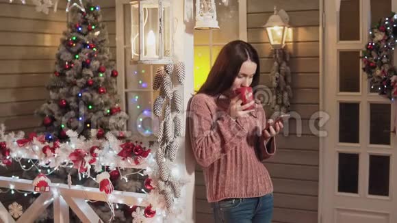 女人微笑着喝茶在门廊用电话背景是圣诞树视频的预览图