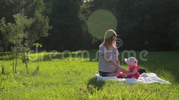 年轻的母亲和她的小女儿坐在绿色的春天草坪上的白色毯子上视频的预览图