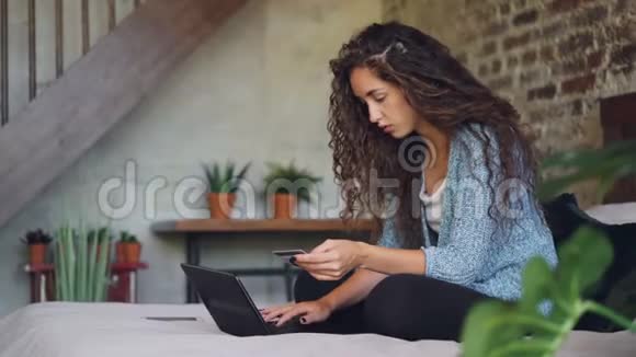年轻的女人拿着银行卡拿着笔记本电脑在家里坐在床上工作网上买东西视频的预览图