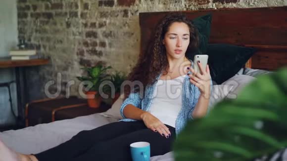 快乐的女孩在网上聊天用智能手机看手机摄像头用微笑坐在卧室的床上视频的预览图