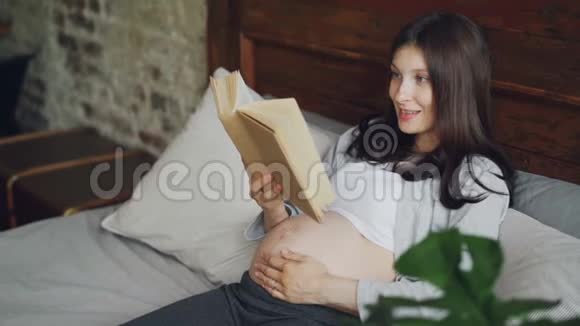 怀孕的年轻女士正在大声读书用爱和温柔抚摸着她的宝宝的肚子微笑的女孩正在享受着视频的预览图