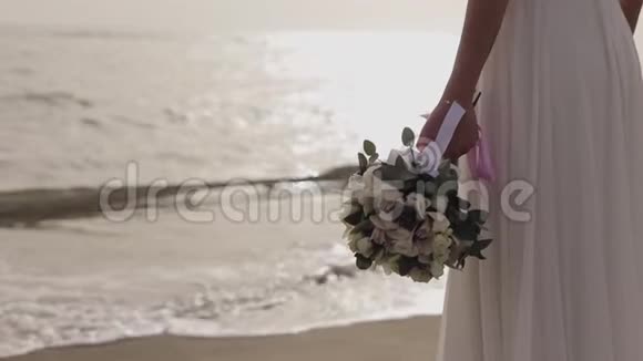 美丽的年轻女孩穿着白色的裙子站在海滩上手里拿着一束花视频的预览图