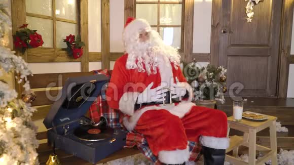 圣诞老人坐在房子附近的圣诞树之间喝牛奶吃饼干听乙烯基的圣诞歌曲视频的预览图