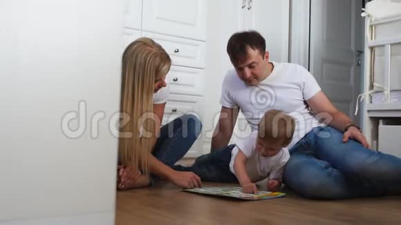 一家三口穿着白色T恤衫和蓝色牛仔裤坐在卧室在卧室的地板上和男孩玩视频的预览图