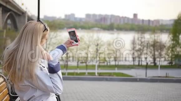 年轻漂亮的金发女郎用智能手机自拍一位年轻女子沿着河堤散步视频的预览图