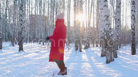 穿着红色冬季夹克和毛毡靴的可爱的年轻女子从寒冷中跳下来并试图在带有桦树的图案城市公园保暖视频的预览图