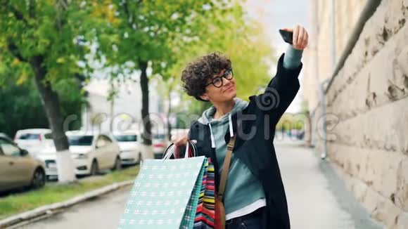 美丽的女孩慢镜头自拍购物袋站在街上拿着智能手机微笑着视频的预览图
