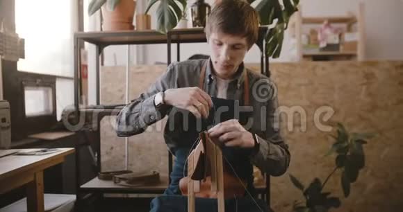 专业男性工匠在轻型现代制造车间用针头在马鞍上缝制手工皮革制品视频的预览图
