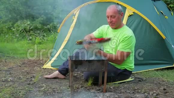 一个穿着绿色T恤的男人坐在森林里靠着绿色的帐篷把烧烤放在烧烤架上总体计划视频的预览图