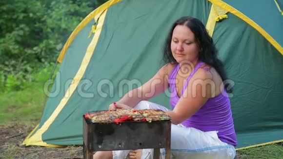 一个穿T恤的年轻女人坐在森林里在绿色的帐篷背景下用她的胸罩准备晚餐视频的预览图