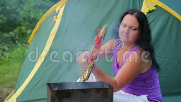 一个穿T恤的年轻女人坐在森林里靠着一个绿色的帐篷在她的烧烤上转动烤肉中间计划莫先生视频的预览图