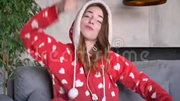 有趣的开朗的女人穿着红色睡衣通过耳机听音乐跳舞坐在沙发上视频的预览图