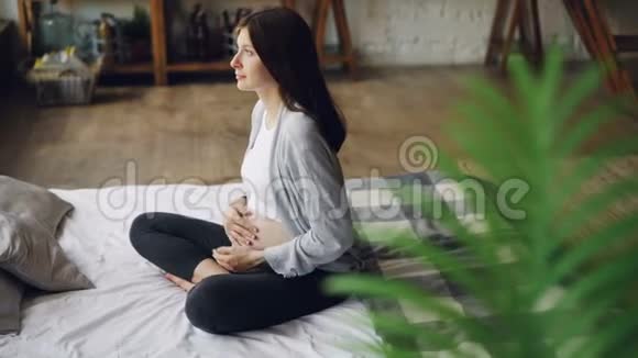 有爱心的怀孕女孩正在抚摸她的婴儿肿块坐在卧室的床上享受怀孕和休息家庭视频的预览图