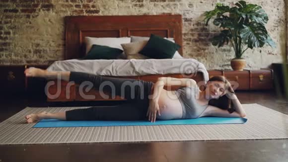 有魅力的年轻女士期待母亲做运动躺在垫子上抬起腿独自在家练习视频的预览图